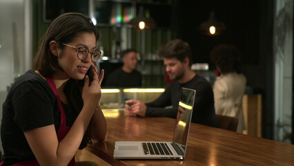 Barista receiving orders on phone browsing online with laptop at cafe. Small business concept of staff using computer at coffee shop