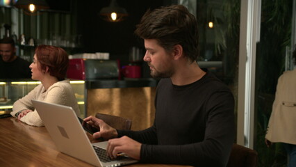 Concentrated man using laptop inside coffee shop checking smartphone device notification. Serious expression looking at computer screen