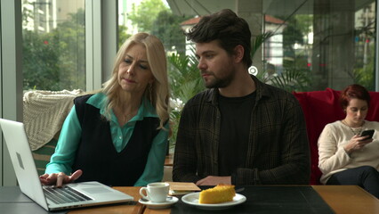 Female leader coaching younger man in front of computer laptop at cafe. Woman explaining protocol to younger staff