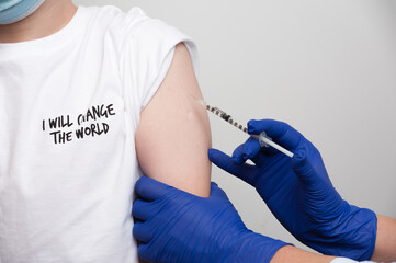 Medical and vaccination topic. Hands of nurse with syringe making vaccine injection to caucasian boy in a white T-shirt with the inscription "I will change the world"