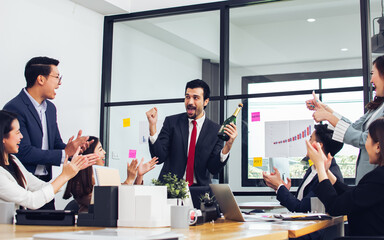Caucasian handsome businessman wearing formal suit, holding bottle of wine to celebrate his success. Asian smiley colleagues clap hands and sitting in meeting room at indoor office or workplace.