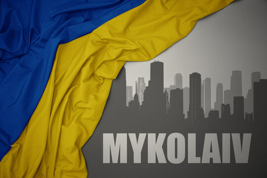 abstract silhouette of the city with text Mykolaiv near waving national flag of ukraine on a gray background.