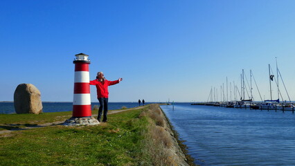 aktiver Senior steht im Dorf Orth in Fehmarn am Hafen mit Segelbooten und dekorativem kleinen Leuchtturm