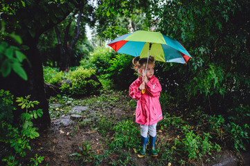 A little beautiful girl blonde, in boots and with umbrella walking along the puddles in the rain. Happy child. A walk under the summers of the rain.
