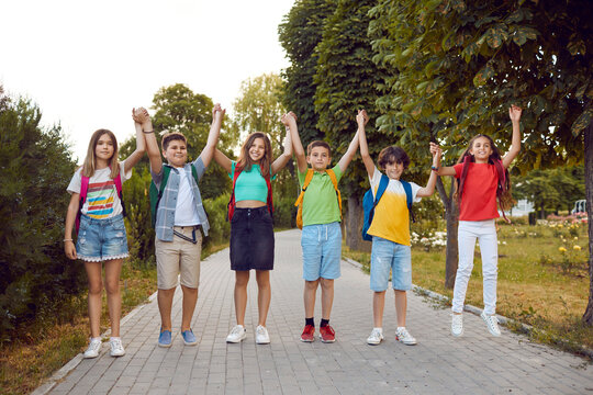 Outdoor group portrait of happy school children. Bunch of joyful school friends in comfortable casual clothes with backpacks standing on a park path, holding hands, smiling and looking at the camera