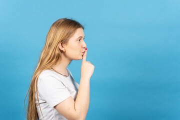 Side view of young blonde woman asking for silence finger on lips isolated on blue background.