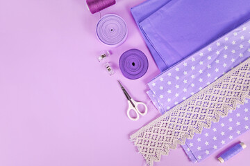 Sewing flat lay with various tools like fabric, scissors, spools and ribbons on violet background