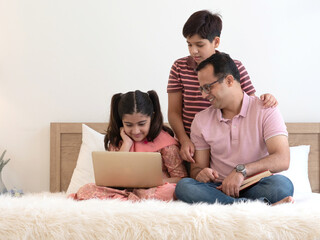 Indian father with two teenage children sitting on the bed together at home using laptop for movie,  homework, online learning. Happy family, single father , education or father's day concept.