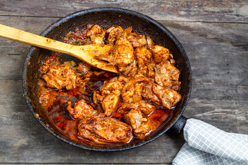 Pieces of baked chicken in sauce in a frying pan with a wooden spatula on a dark table.