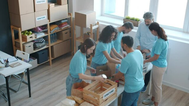 Diverse group of enthusiastic volunteers packing donation boxes with food and talking in charity fund office. Teamwork and humanitarian aid concept.