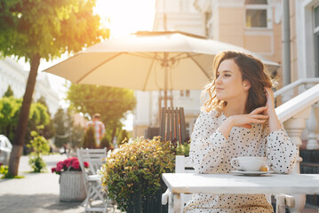 Charming young lady sitting at coffee table in street cafe and touching her curly hairstyle outdoors. Posing cute woman in dress enjoying summer sunny weather during coffee break, lifestyle