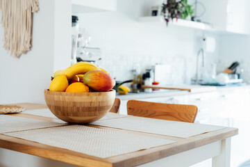Home kitchen counter table with selective focus on bamboo bowl with exotic fruits on it with blurred background of modern cozy white kitchen. Home interior design details. Copy space.