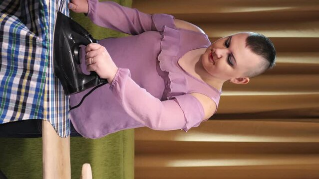 A woman with a shaved mohawk on her head irons linen on an ironing board at home. Vertical video