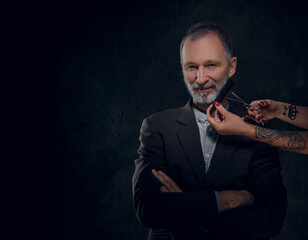 Shot of female hands cutting beard of senior man dressed in elegant suit against dark background.