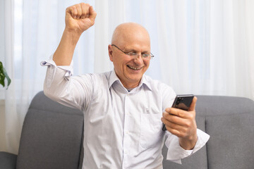 Happy excited grandfather holding smartphone, laughing, shouting for joy, getting good news, reading text message on screen, making winner yes hand gesture. Surprised older man winning prize