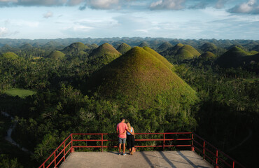 A young couple of travelers enjoy panoramic views at sunset in the Chocolate Hills of Bohol,...