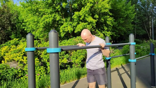 Young man trains on outdoor sports field in city park. Man does gymnastics on parallel bars. Training of muscles of biceps and triceps. Handsome bald man push-ups.  Healthy lifestyle