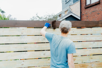 Young man worker paints with a roller a wooden board fence in the garden. DIY, Do it yourself concept. House improvement. Home renovation and refurbishment. Selective focus, copy space.
