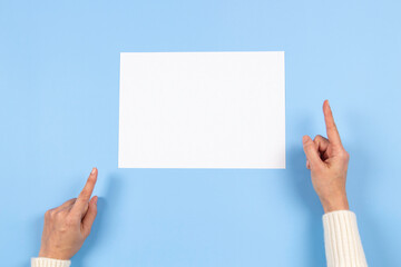 Female hands pointing to blank white paper sheet on light blue background. Top view. Mockup paper...