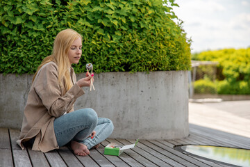 Young beautiful blond woman eating sushi outdoors, on the wooden terrace, by modern building in the...