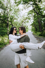 Fototapeta premium A bearded long-haired stylish man holds his beloved hippie brunette woman in sunglasses in his arms and circles her in nature in the park. Portrait, photo of newlyweds, concept.