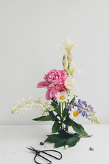 Modern summer flowers composition on rustic white table indoors. Creative floral image. Stylish peony, lupin,iris and daisy arrangement on kenzan with scissors on rustic wood.