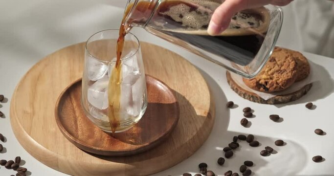 4k video, pouring brewing coffee into glass full of ice is served with cookies on white table. Making cold coffee drink in slow motion. 