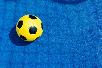 Fototapeta na wymiar Sports court background. Light blue field rubber ground with shadow from football goal net and yellow soccer ball on sunny day outdoors. Top view