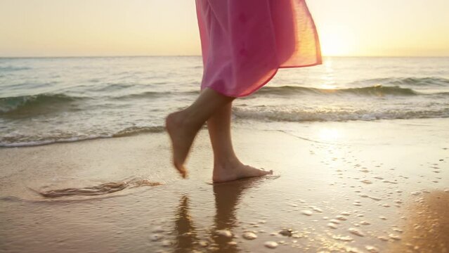 Slow motion woman feet walking barefoot by beach at golden sunset leaving footprints in wet sand. Female tourist on summer vacation walking in golden liquid by ocean shore on Hawaii island at sunset