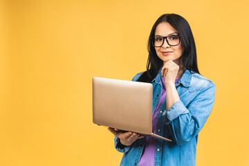 Portrait of happy young beautiful surprised woman with glasses standing with laptop isolated on yellow background. Space for text. - 512674404
