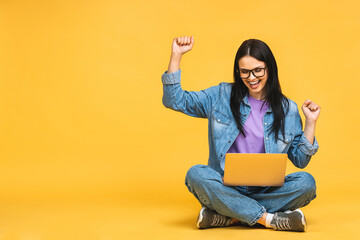 Happy winner! Business concept. Portrait of happy young woman in casual sitting on floor in lotus pose and holding laptop isolated over yellow background. - 512674439