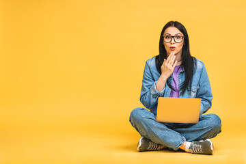 Business concept. Portrait of amazed shocked surprised young woman in casual sitting on floor in lotus pose and holding laptop isolated over yellow background.