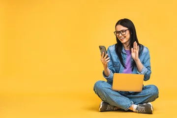 Foto op Plexiglas Business concept. Portrait of happy young woman in casual sitting on floor in lotus pose and holding laptop isolated over yellow background. Using mobile phone. © denis_vermenko