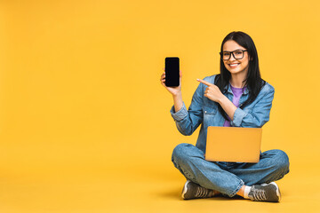Business concept. Portrait of happy young woman in casual sitting on floor in lotus pose and holding laptop isolated over yellow background. Using mobile phone. Showing phone screen. - 512674486