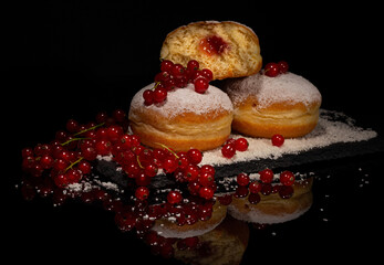 Berlinere German sweet donuts sprinkled with powdered sugar filled with fruit jam surrounded by ripe berries on a black slate board on a black background with reflection