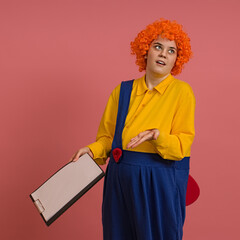 funny clown in a wig and a yellow-blue suit is perplexed and holds a clipboard in his hand on a colored background