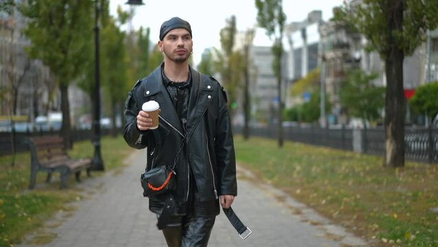 Dolly shot of confident positive gay man strolling on summer autumn alley in city drinking coffee looking around. Portrait of stylish handsome Caucasian LGBTQ person enjoying weekend outdoors leaving