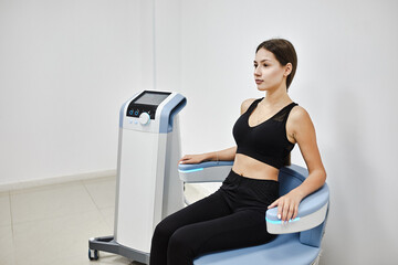 Young woman sitting on electromagnetic chair for stimulation of deep pelvic floor muscles and restoring neuromuscular control at the clinic - 512677634