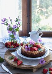 Tavola apparecchiata con una torta di fragole, una ciotola piena di frutta e un mazzo di fiori