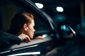 a close horizontal photo from the side, at night, of a woman sitting in a black car and looking out of the window looking into the side view mirror