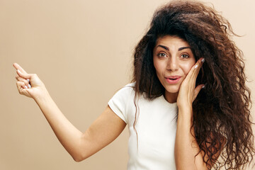 SEASONAL SALE OFFER. Excited beautiful Latin female in white t-shirt with afro say Look at this, look at camera, show copy space, free place for design ad. Fashion. Studio shoot over beige background