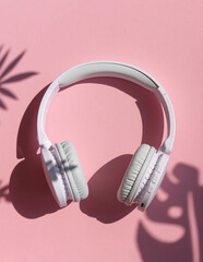 Wireless white headphones with tropical leaves shadows background on the pink close up. Listening to summer music concept	
