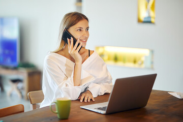 Beautiful woman in white shirt is having a phone call at home with a laptop and cup of coffee at the table.