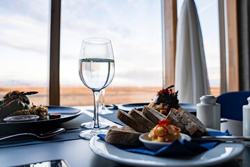 Close-up of wineglasses and tasty food on dining table. Gourmet meal is served against window with view of sky. Concept of beautiful catering in restaurant at luxurious hotel.