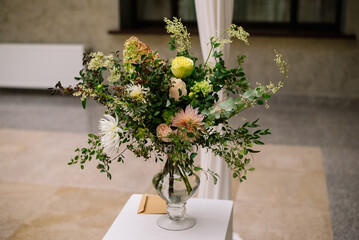 Element of wedding decor or celebration event. A beautiful bouquet in pastel colors stands on the table in a glass vase next to the envelope with the postcard. High quality photo