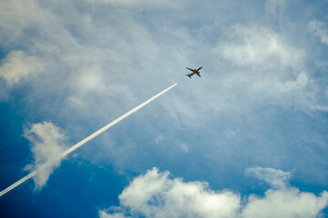 Small dark airplane silhouette flying high up in the blue cloudy sky. The aircraft in the sky. Airplane condensation trace. Traveling by plane.