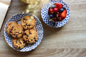 Plate of chocolate chip cookies, cup filled with strawberries, blueberries and cherries, open book and vase with gypsophila flowers on the table. Flat lay.