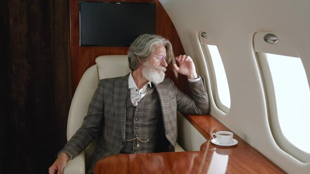 Stylish mature millionaire man wearing classical suit luxury flying in private airplane. Confident CEO businessman looking through corporate jet window. Luxury traveling in private jet.