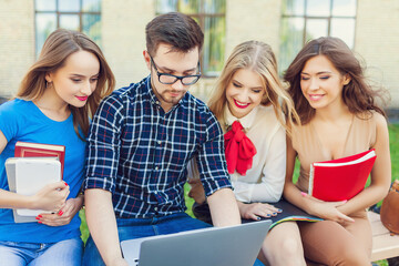 
A group of cheerful students near the university building on the lawn with a laptop, books and notebooks