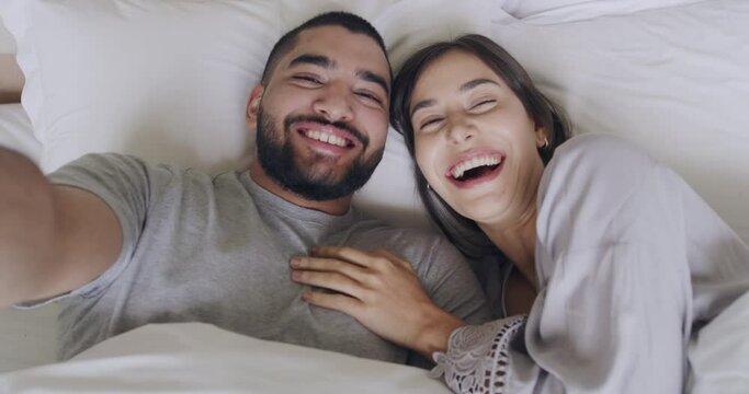 Portrait of a young latin couple kissing while taking selfies in bed together. Loving boyfriend taking a photo while giving his girlfriend a kiss. Cheerful husband and wife lying in bed in the morning